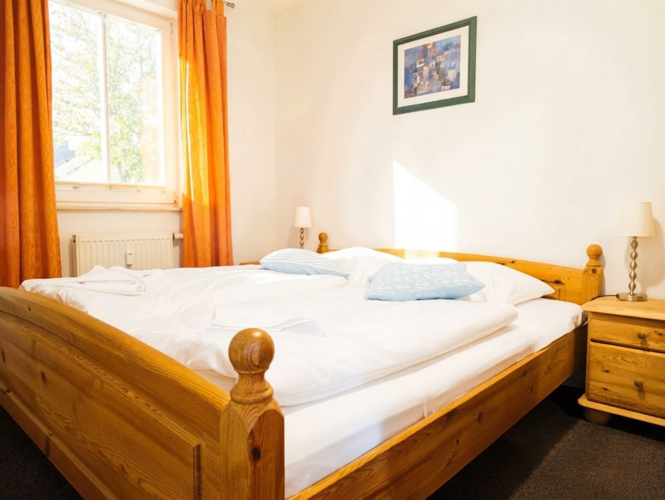 Uma cama de madeira com roupa de cama branca, uma mesinha de cabeceira com abajur e uma janela com cortinas.