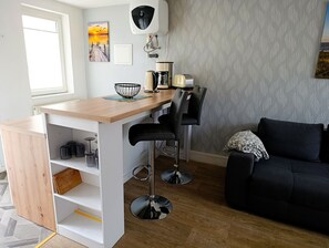 A compact living space with a kitchenette, a dining area with bar stools, a black sofa, and a patterned wall. - Apartment Rosenhof TW 102 - TW 102 (Kühlungsborn)