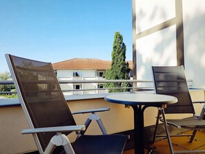 Varanda com duas cadeiras e uma mesa pequena, com vista para um edifício com telhado de azulejos e uma árvore.