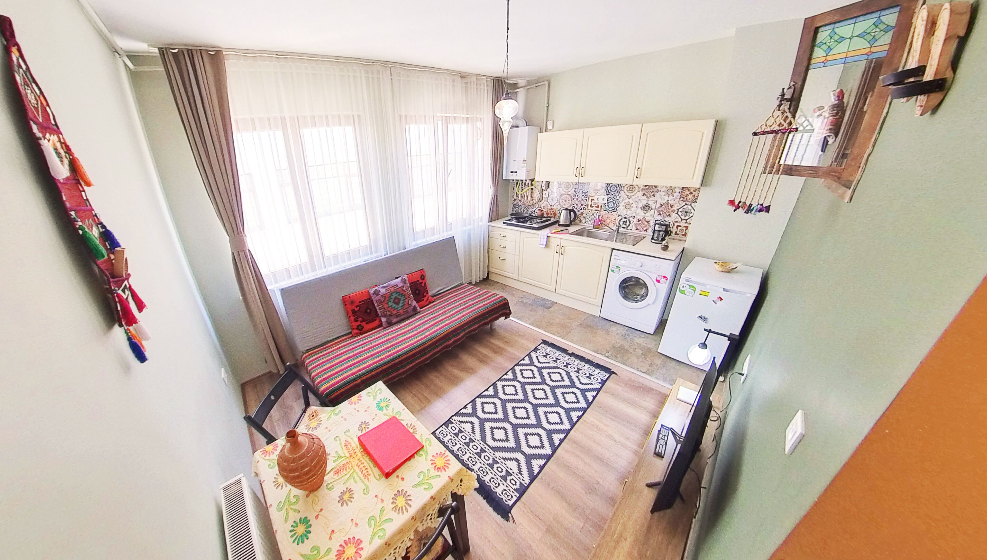 A compact living space with a sofa, a dining table, a kitchen area including a washing machine, and a small window with curtains.