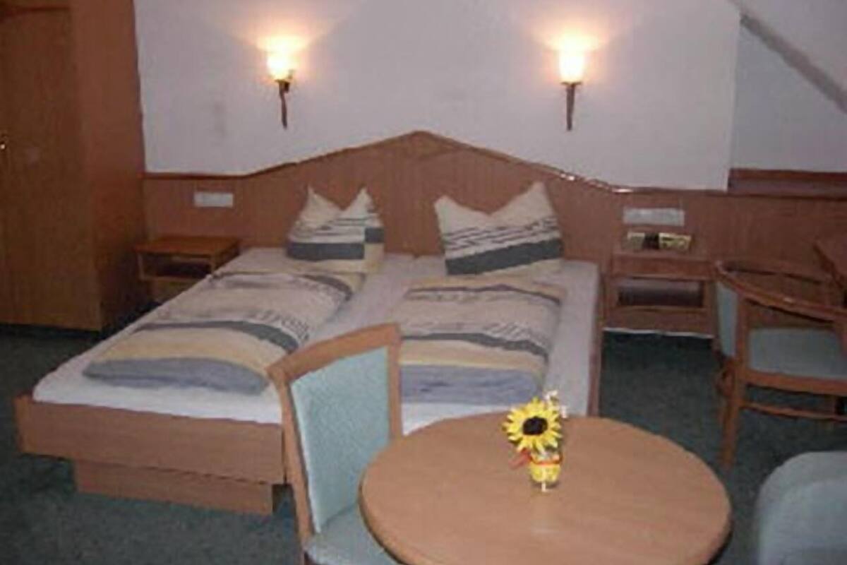 Una cama doble con cabecera de madera, dos mesitas de noche, una pequeña mesa redonda con una decoración de girasol y dos sillas.