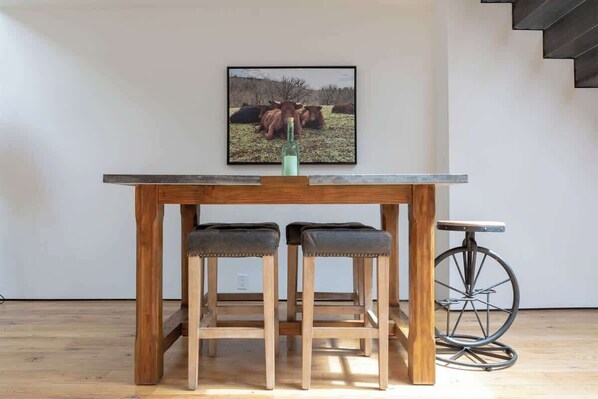 Una mesa de madera para comer con dos taburetes, una botella y un cuadro con vacas en la pared.