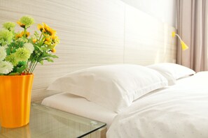 A hotel room with a bed, white pillows, and a glass table with a yellow vase of flowers.