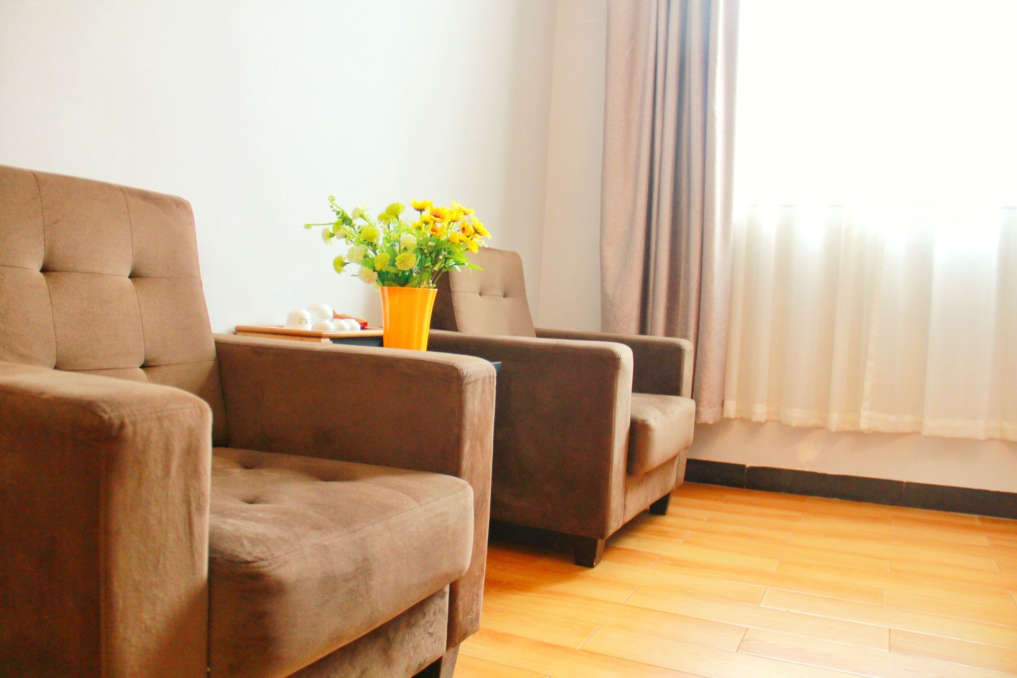 Una sala de estar con dos sofás marrones, una mesita con un jarrón de flores amarillas y una ventana con cortinas.