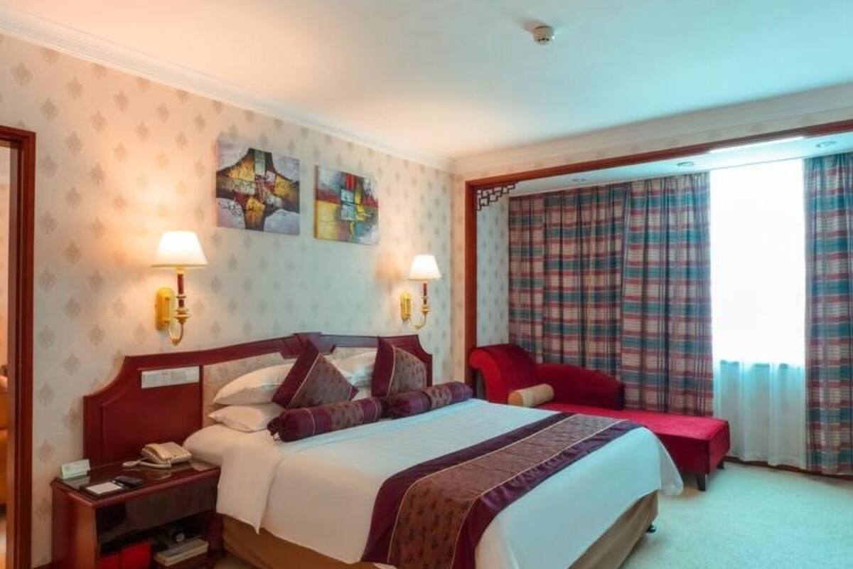 Una habitación de hotel con una cama grande, dos mesitas de noche, un sillón rojo y una ventana con cortinas escocesas.