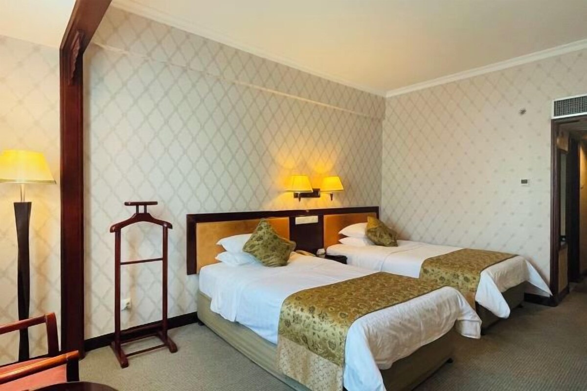 A hotel room with two beds, a wooden chair, a coat rack, and a wall-mounted lamp.