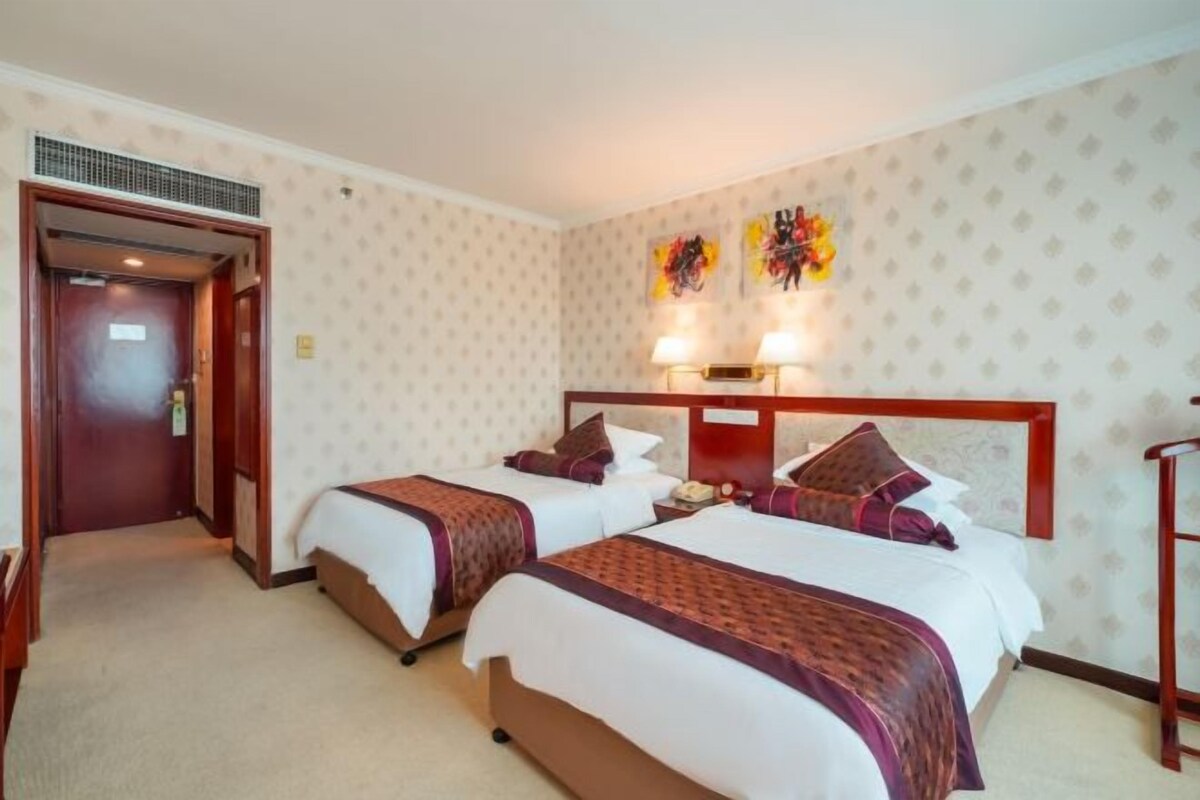 A hotel room with two beds, a wooden headboard, a nightstand, and a wall-mounted lamp.