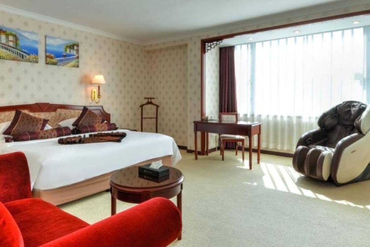Habitación de hotel con cama, un sillón rojo, una silla de masaje, una mesita y una ventana con cortinas.