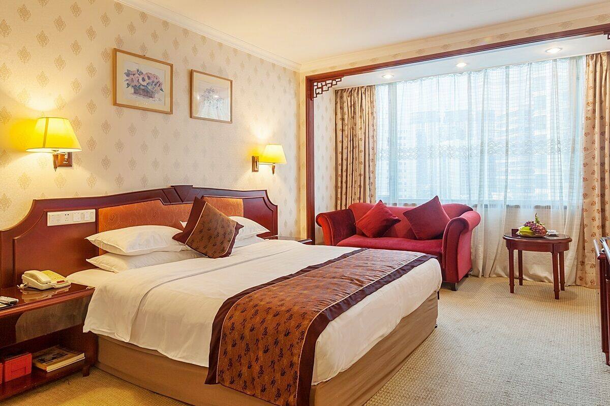 Una habitación de hotel con una cama grande, dos lámparas de noche, un sillón rojo, una mesita con fruta y una ventana con cortinas.