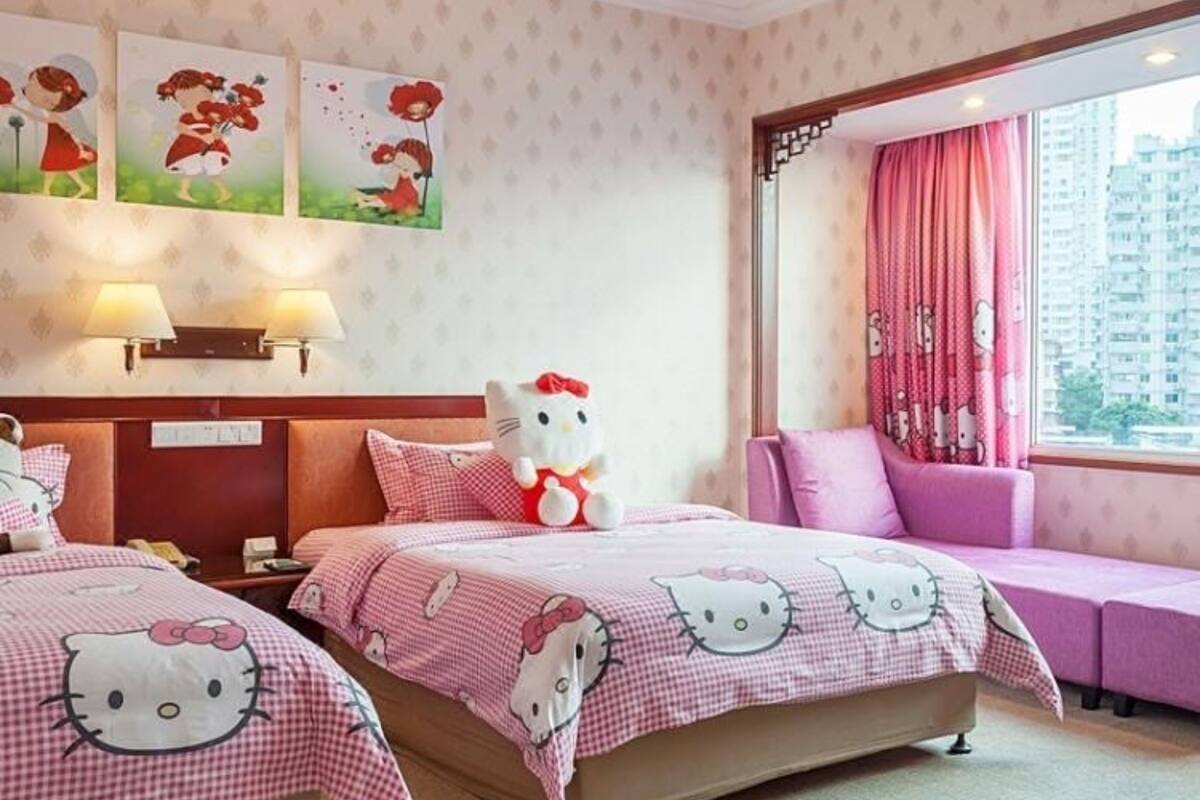 Habitación de hotel con dos camas, cada una con ropa de cama de Hello Kitty, un peluche grande de Hello Kitty y un sofá rosa. Hay lámparas fijadas a la pared y una ventana con cortinas rosas.