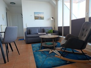 Un salon moderne avec un canapé gris, un fauteuil à bascule, une petite table sur laquelle est posée une plante, et un tableau accroché au mur.