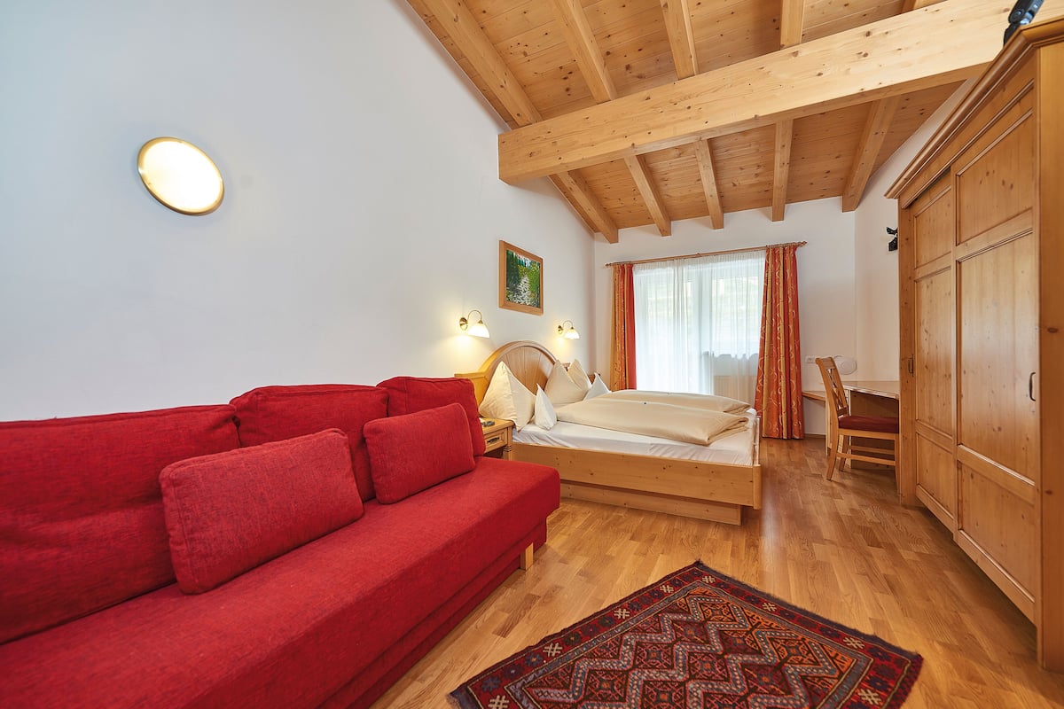 A room with a wooden ceiling, a red sofa, a bed with a wooden frame, a wooden wardrobe, and a patterned rug.