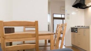 Una sala de estar con sofá, una mesa de comedor de madera con sillas, una zona de cocina con estufa y armarios, y un reloj en la pared.