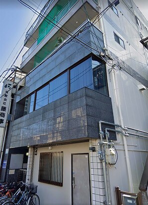 Two-story building with a flat roof and balcony, featuring large windows and a door on the ground floor.
