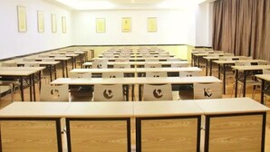 Een congrescentrum met rijen bureaus en stoelen, een podium aan de voorkant, en ingelijste foto's aan de muren.
