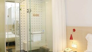 Een hotelkamer met een glazen douchecabine, een bed met witte lakens, een nachtkastje met een rode lamp en een geruit vloertapijt.