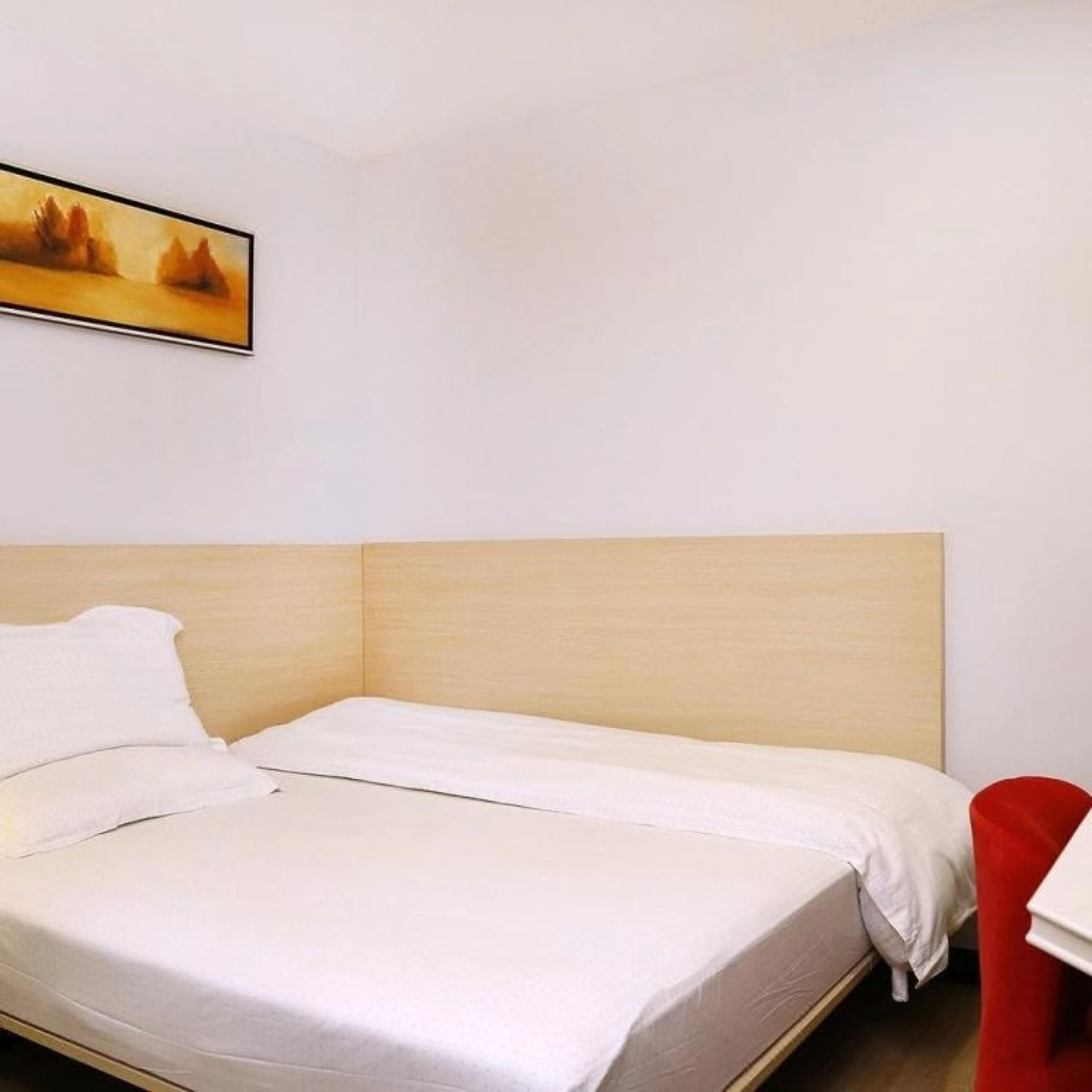 Una cama individual con cabecera de madera, ropa de cama blanca y una silla roja en una habitación con paredes blancas.