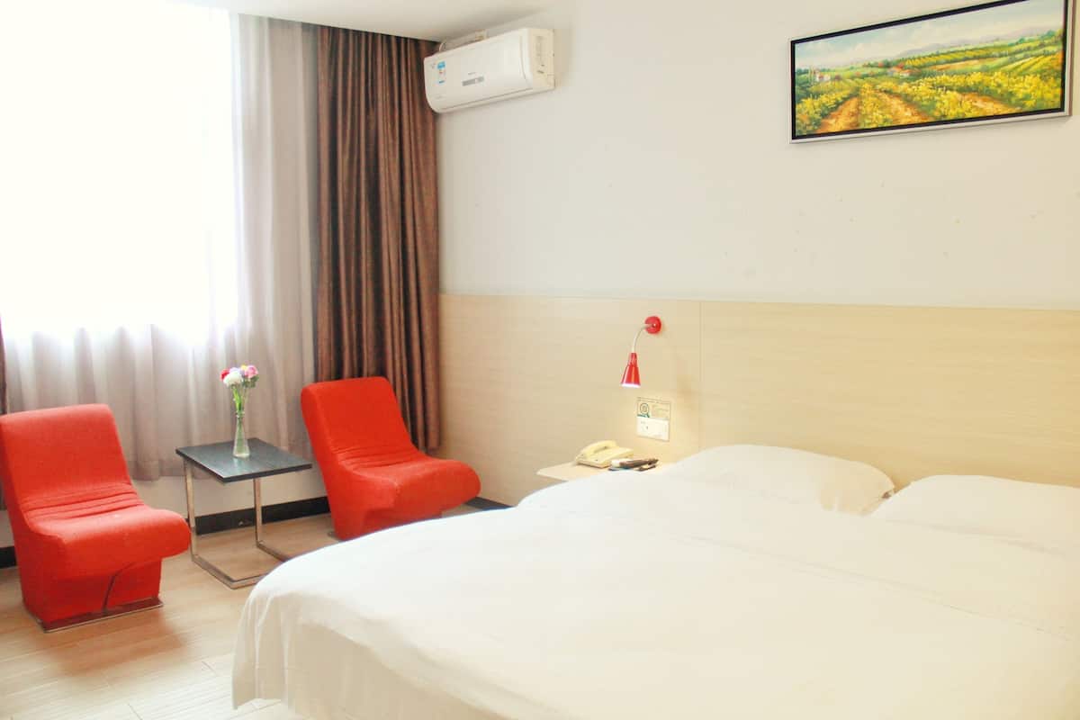 Une chambre d’hôtel avec un lit, deux fauteuils rouges, une petite table sur laquelle est posé un vase de fleurs, une table de chevet avec une lampe, et un tableau accroché au mur.