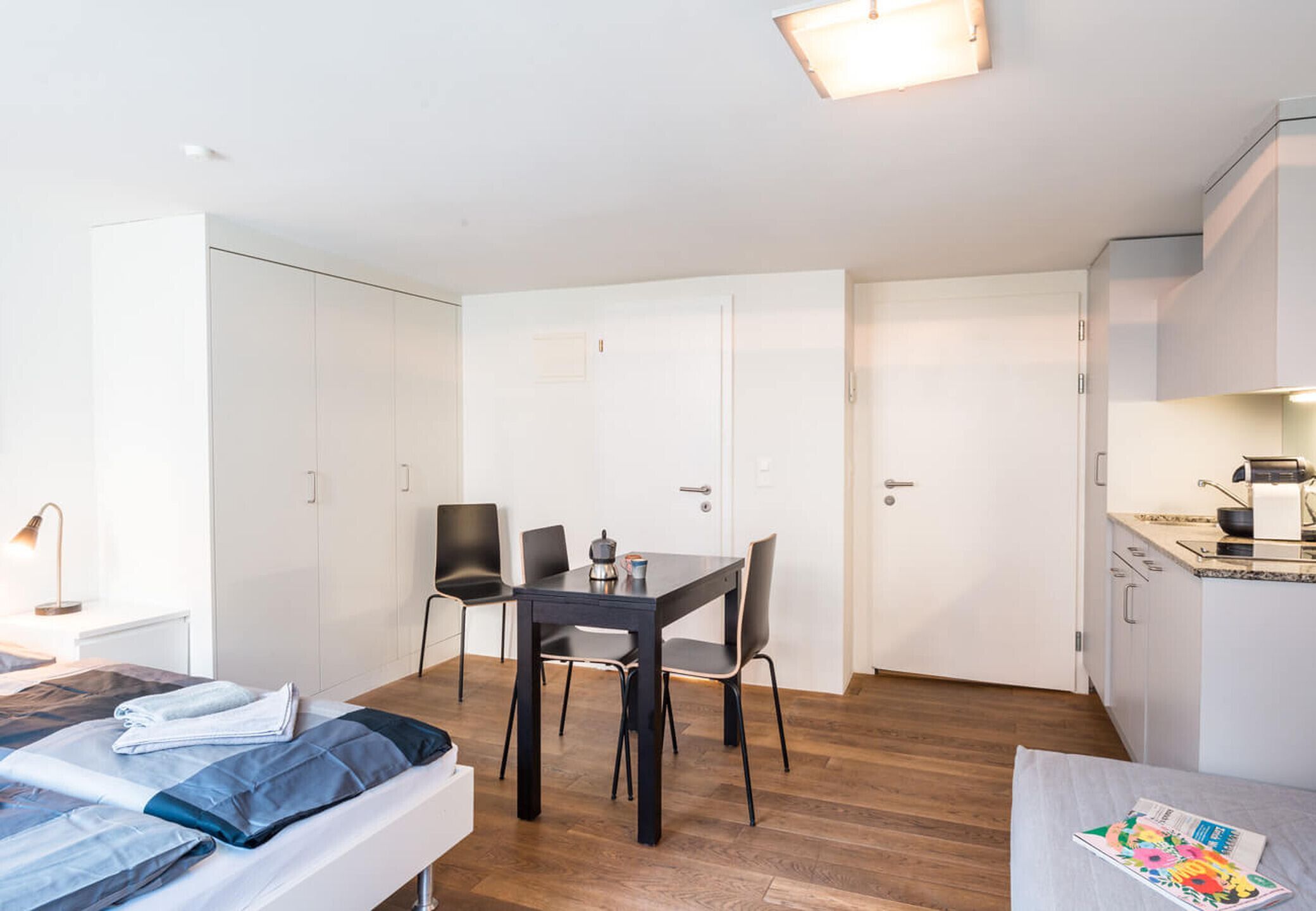 Una habitación compacta con una cama, una pequeña mesa de comedor con sillas, una kitchenette y armarios blancos.