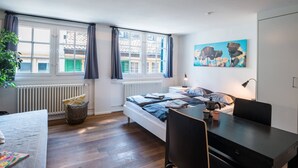 A bedroom with a bed, a desk, a chair, a radiator, a window with curtains, and a painting on the wall. - ZH Schmidgasse I - Hitrental Apartment (Zürich)