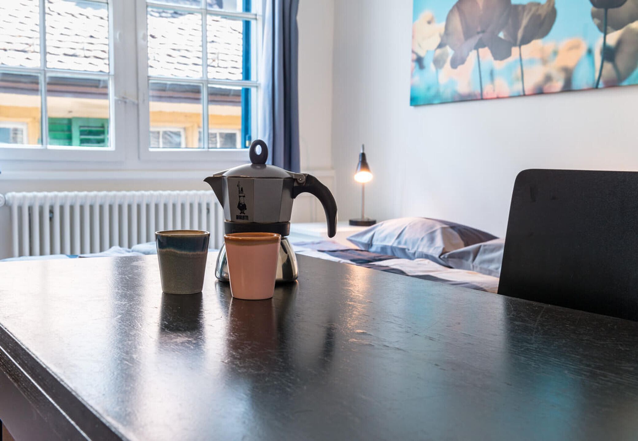 Una zona de cocina con una tetera y dos tazas sobre una mesa, una ventana con persianas y un cuadro en la pared.