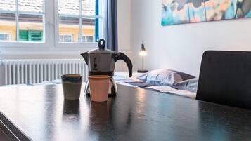 Una zona de cocina con una tetera y dos tazas sobre una mesa, una ventana con persianas y un cuadro en la pared.