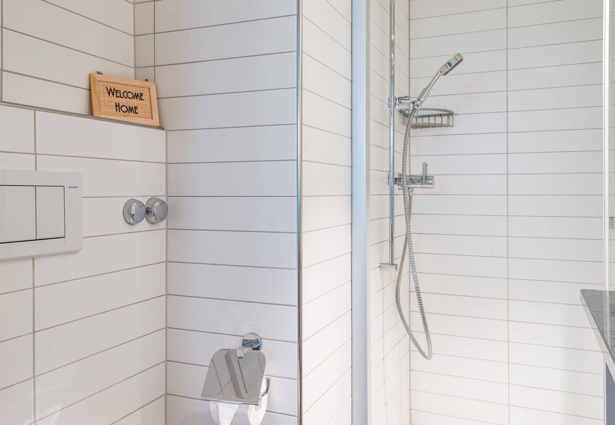 Un baño con paredes de azulejos blancos, ducha con cabezal de ducha de mano, toallero y cartel de bienvenida.