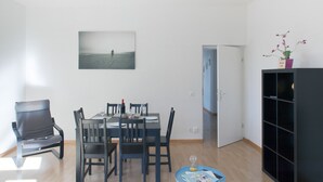 A modern living room with a dining area, featuring a table set for four, a black bookshelf, and a painting of a beach scene. - ZH Seefeld - Hitrental Apartment (Zürich)