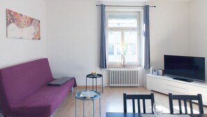 A modern living room with a purple sofa, a dining table with chairs, a flat-screen TV, and a window with curtains. - ZH Seefeld - Hitrental Apartment (Zürich)