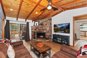 A cozy living room with a stone fireplace, leather couches, a wooden coffee table, and a TV mounted on the wall.