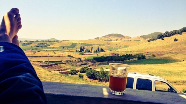 Une personne est assise sur une bordure, un verre à la main, et contemple un paysage rural avec des champs et des collines.