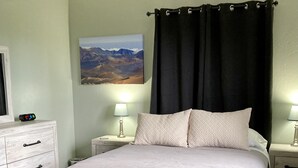 Un dormitorio ordenado con cama, dos lámparas de noche, una mesita de noche, un banco con toallas, un cuadro en la pared y un ventilador de techo.