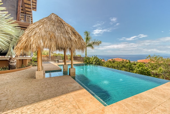 A swimming pool with a thatched roof structure, surrounded by stone tiles and palm trees, overlooking a coastal landscape. - Beautiful condo w/breathtaking ocean & mountain views and shared pool (Playa Ocotal)