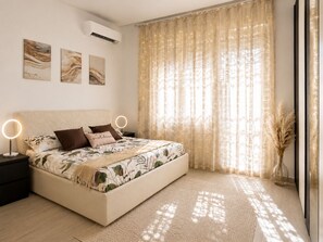 Un dormitorio con cama, mesitas de noche, aire acondicionado y una ventana con cortinas traslúcidas.
