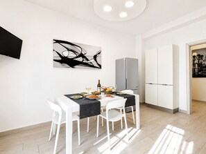 Un espace repas moderne avec une table dressée pour quatre personnes, des chaises blanches, un tableau abstrait noir, un réfrigérateur et un meuble de rangement.
