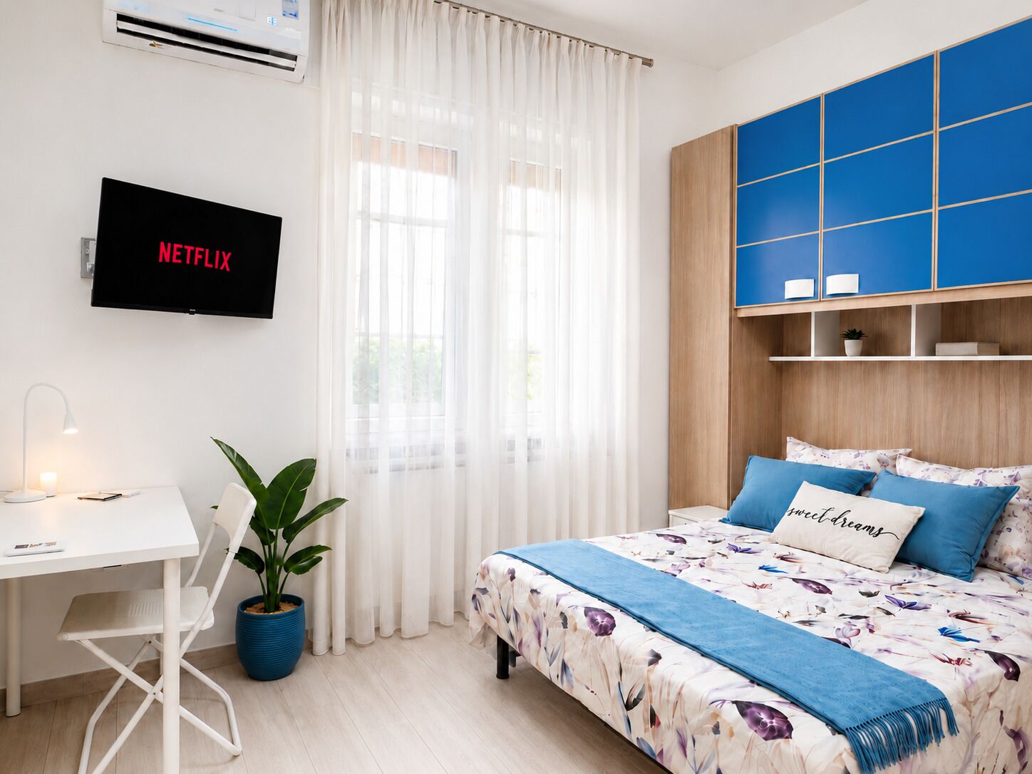 Quarto com cama, escrivaninha, planta em vaso, televisão e janela com cortinas.