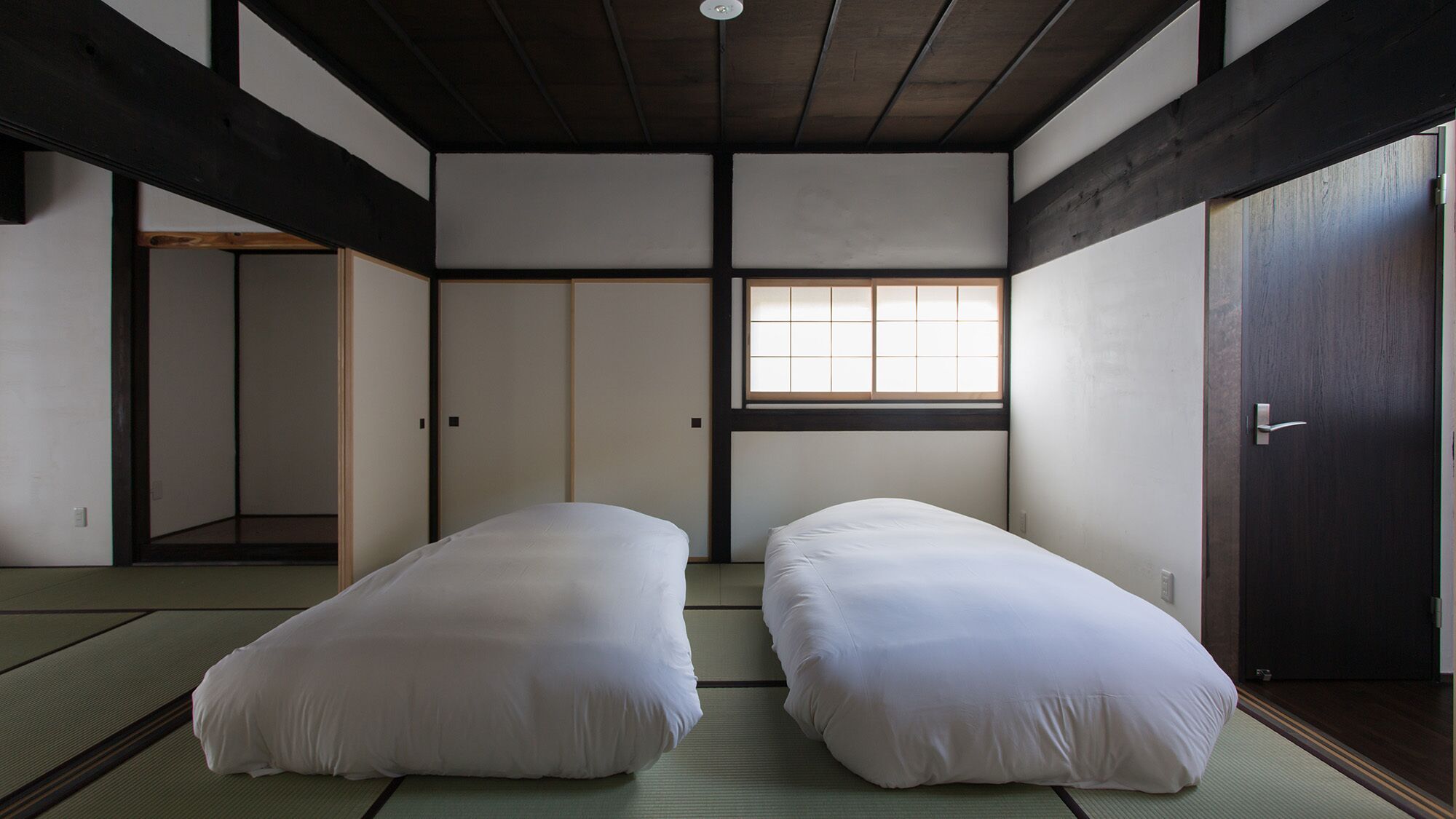 Una habitación japonesa tradicional con suelo de tatami, dos camas individuales, una puerta corredera y una ventana con diseño de cuadrícula.