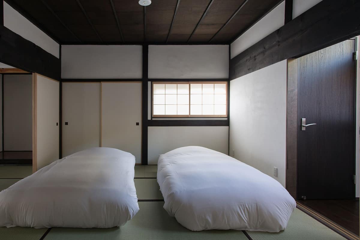 Una habitación japonesa tradicional con suelo de tatami, dos camas individuales, una puerta corredera y una ventana con diseño de cuadrícula.