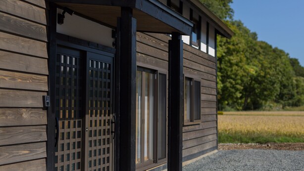 A wooden building with a dark door and windows, set against a backdrop of a field and trees. - Solii (Ishikari)