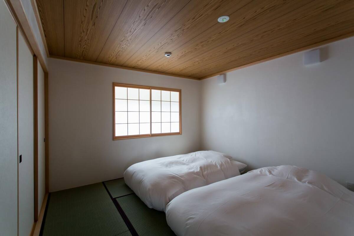 Una habitación con techo de madera, ventana con pantalla shoji, esteras de tatami y dos camas.