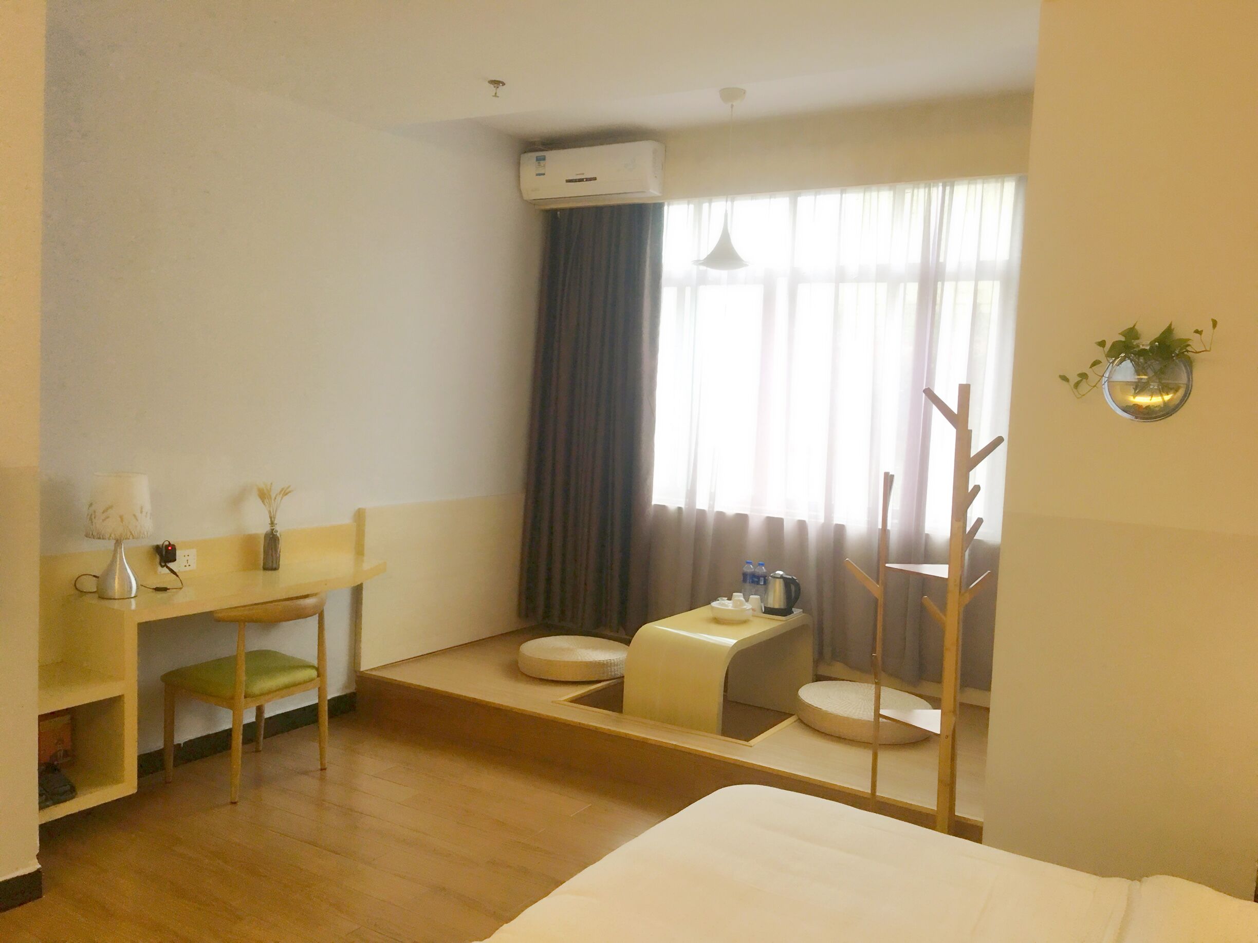 A hotel room with a bed, a desk with a chair, a small table with a kettle, a toilet, and a window with curtains.
