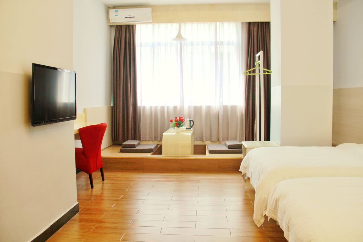 Hotelkamer met een bed, een televisie, een rode stoel, een tafeltje met een plant en een raam met gordijnen.