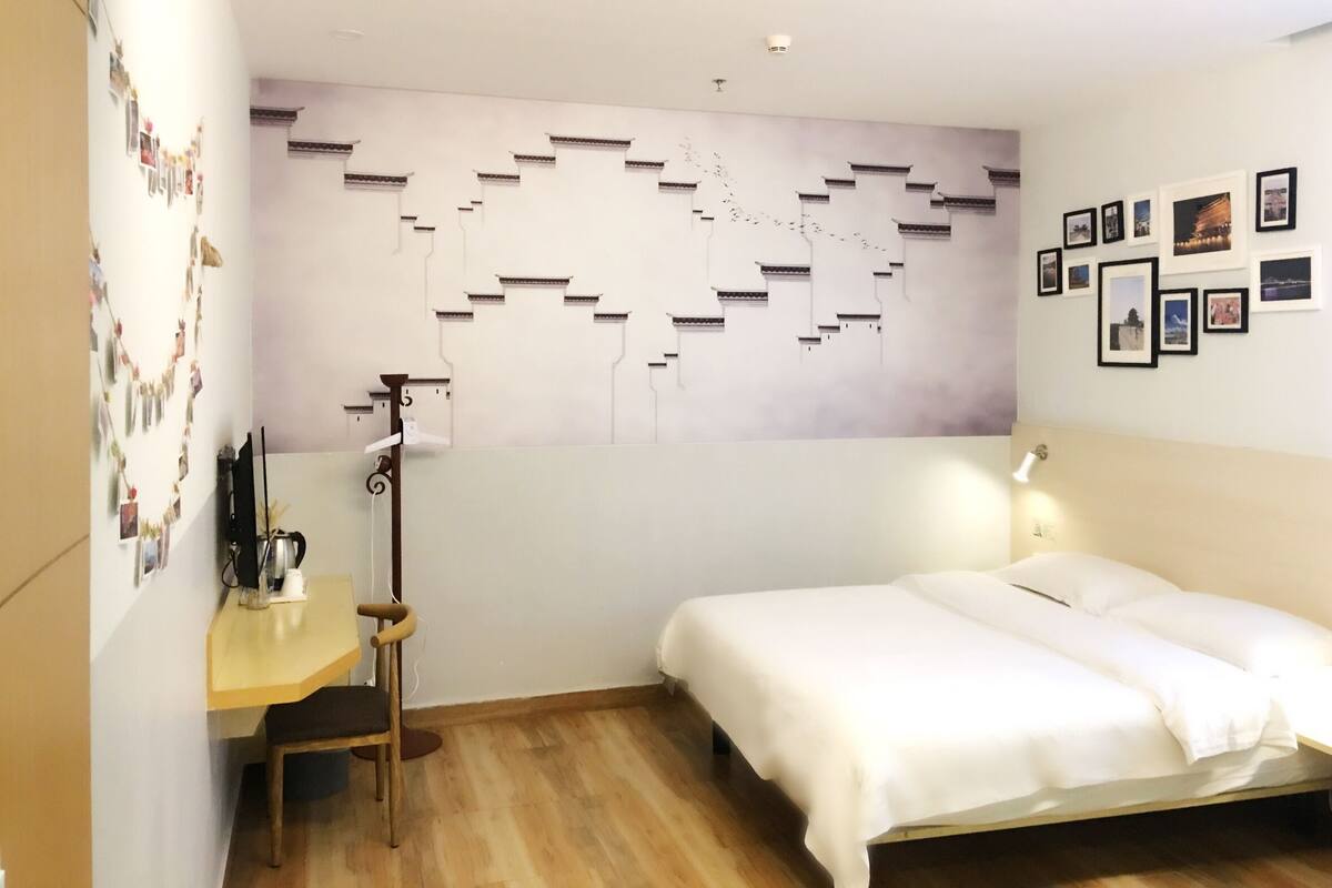 Een slaapkamer met een bed, een bureau en een stoel. Er is een wand met foto’s en een decoratieve wandversiering.