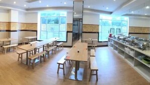Un vaste espace dédié à la restauration, avec de nombreuses tables et bancs en bois, de grandes fenêtres laissant entrer la lumière naturelle et un buffet installé sur le côté droit.