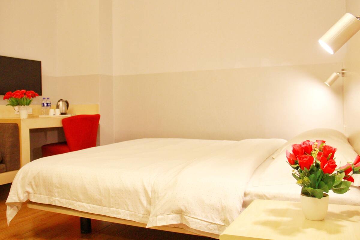 Hotelkamer met een bed, een bureau, een stoel, een kleine tafel met een bloevas en een tv aan de muur.