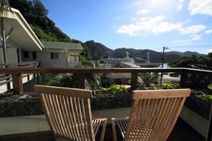 A balcony with two wooden chairs, a stone wall, and a view of a bridge and mountains.