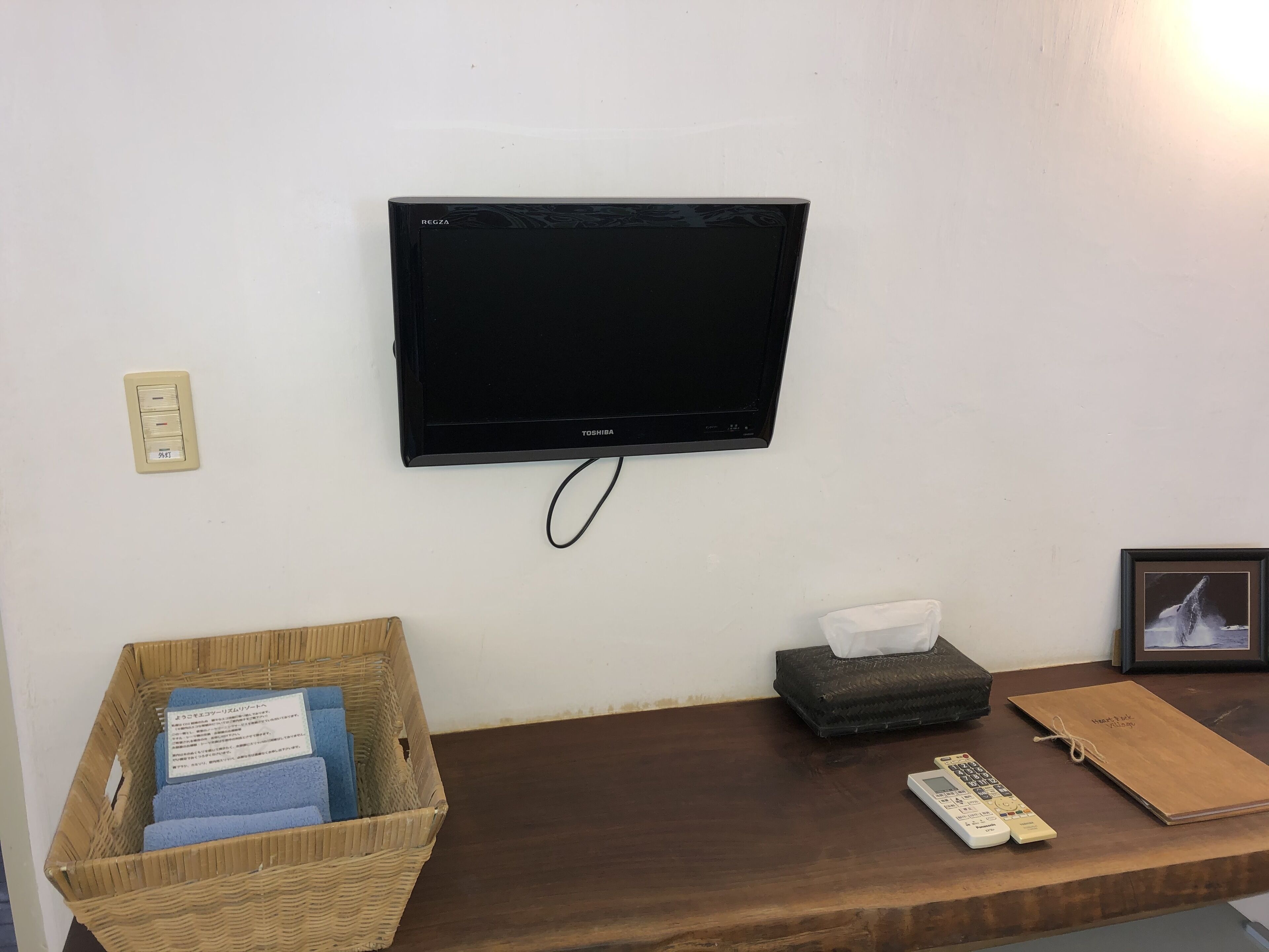 Une pièce avec un téléviseur fixé au mur, un panier en osier contenant des serviettes bleues, une boîte de mouchoirs, une télécommande et un tableau encadré posé sur une table en bois.