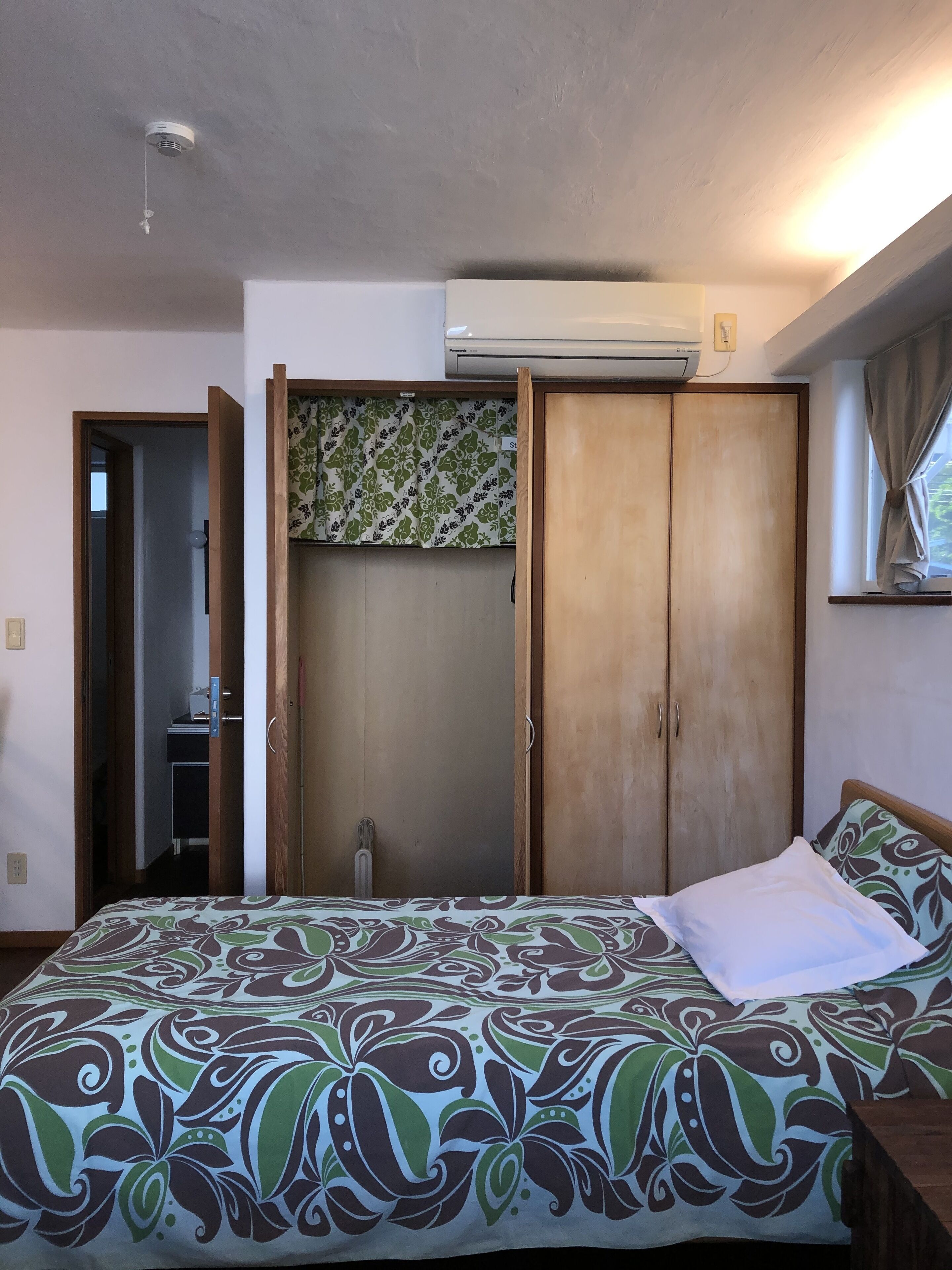 A bedroom with a bed, a wardrobe, an air conditioning unit, and a window with curtains.