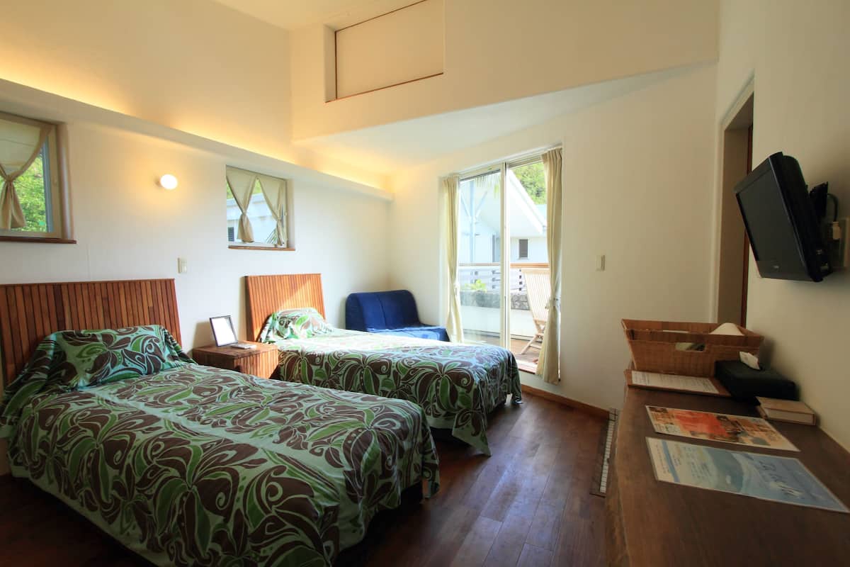 Ein Hotelzimmer mit zwei Betten, einem Schreibtisch, einem Stuhl, einem Fernseher und einem Balkon mit Vorhängen.