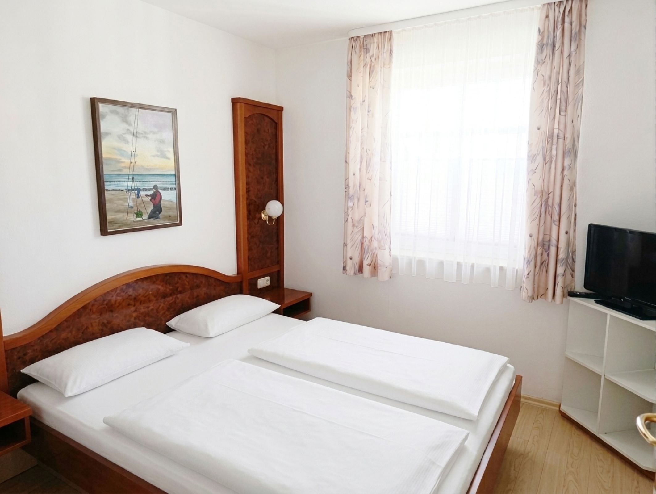 A double bed with white linens, a wooden headboard, a nightstand, a wooden wardrobe, a television on a stand, and a window with sheer curtains.